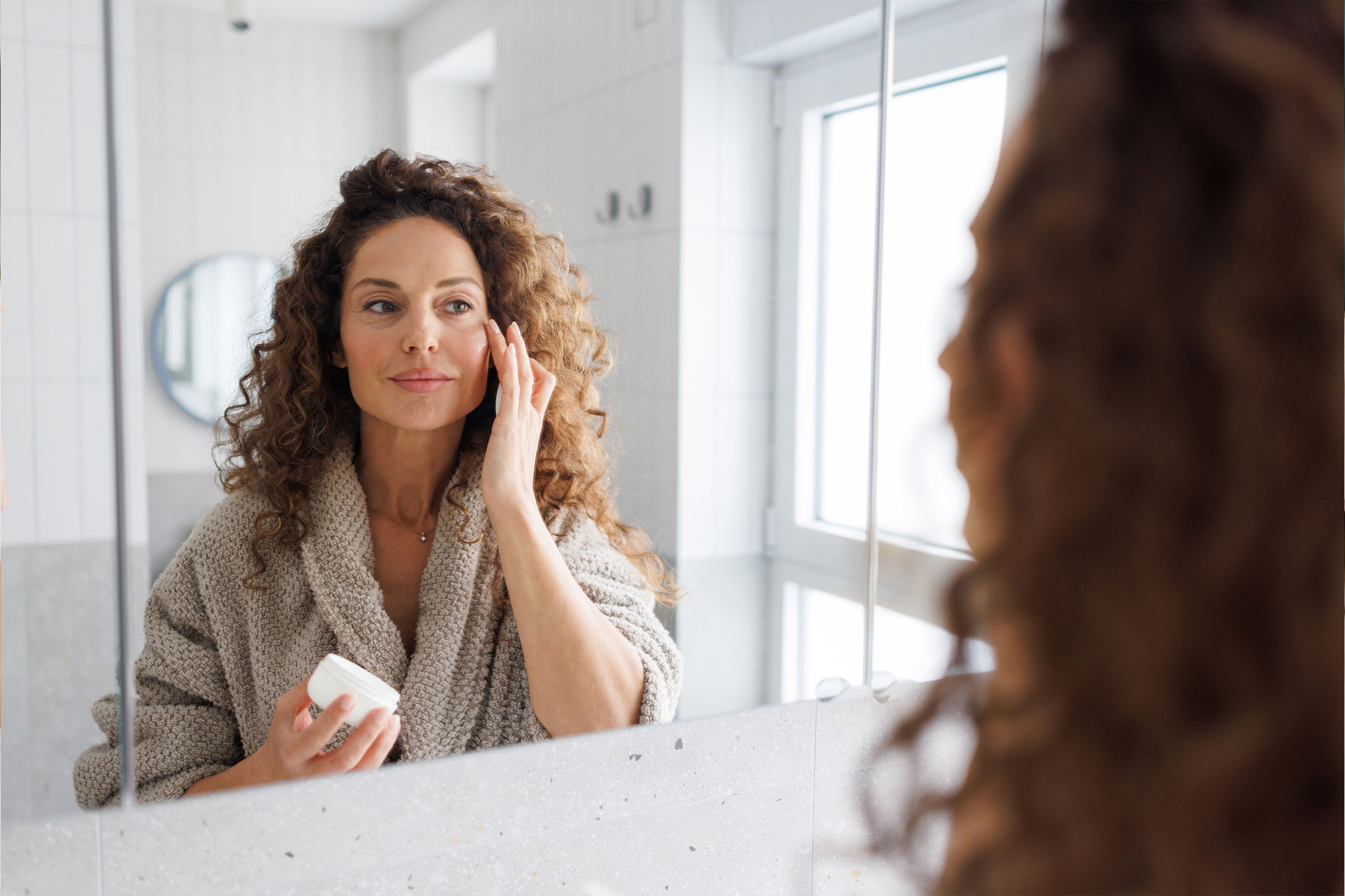A woman applying facial cream in front of a bathroom mirror.
