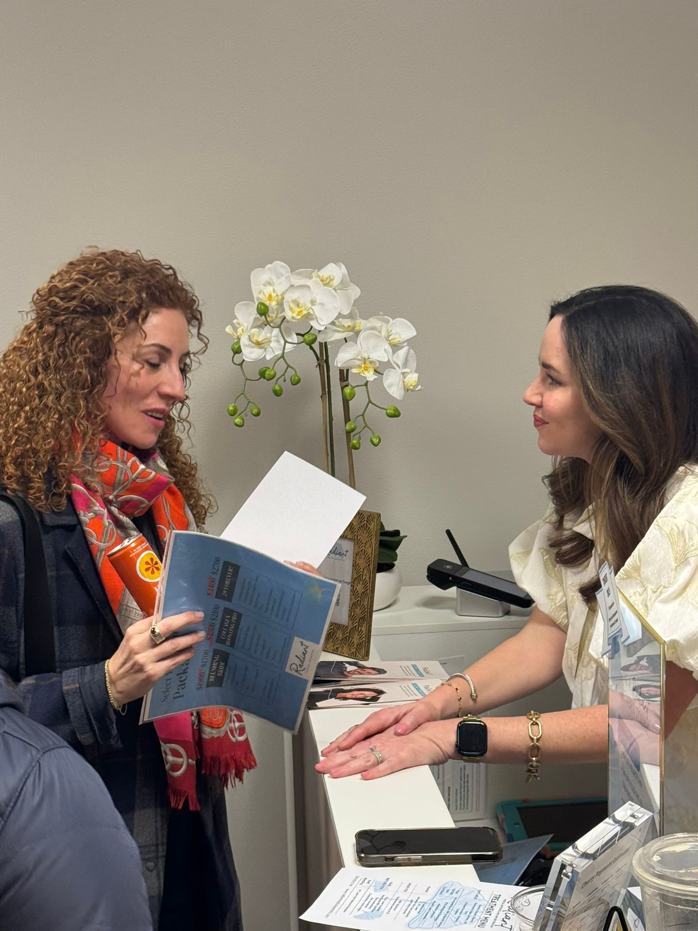 A woman in a scarf shows a document to another woman at a reception desk with a white orchid.