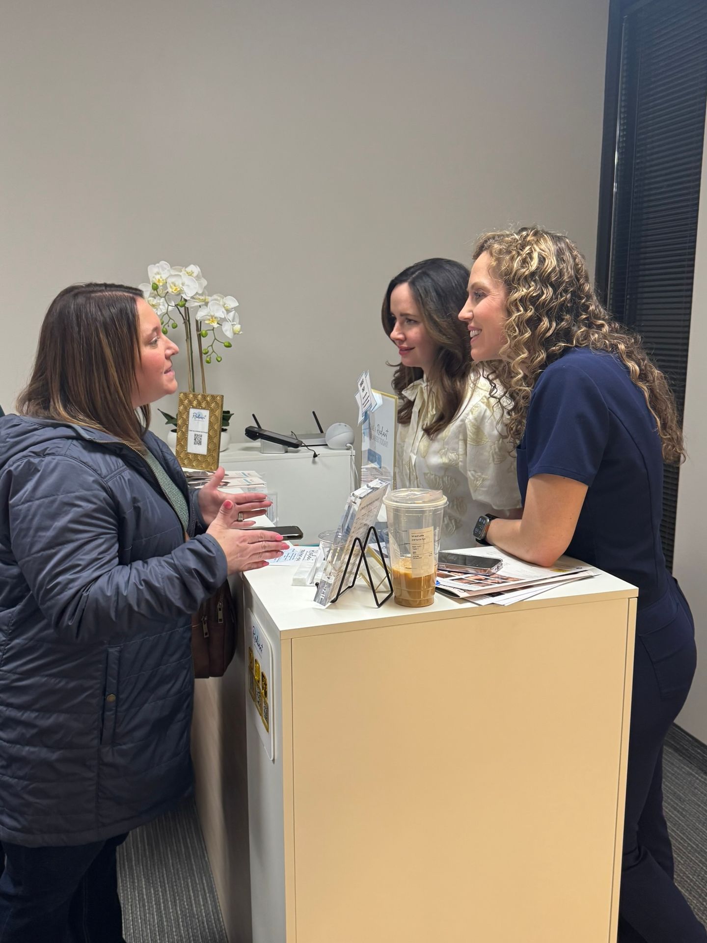 Three women engaged in conversation at a reception desk with flowers and paperwork.