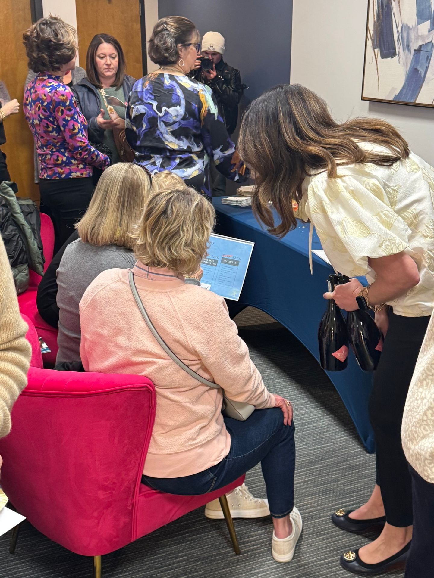 People gathered around a table, one person seated viewing a laptop, others standing and holding items.