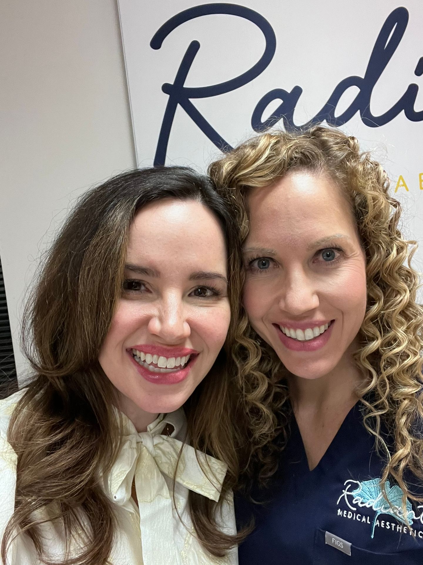 Two smiling women posing in front of a Radiesse sign at a medical aesthetics clinic.