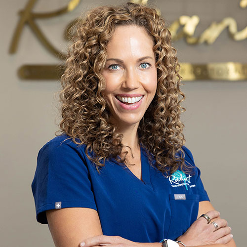 A smiling woman in blue medical scrubs with curly hair stands in front of a wall sign.