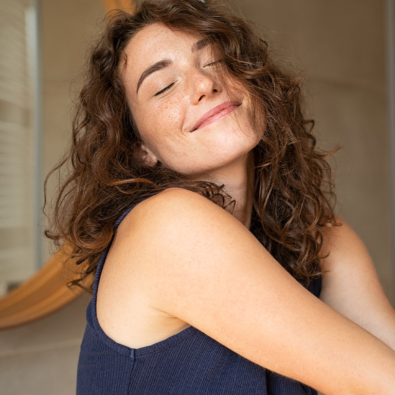 A woman with curly hair and a content expression, eyes closed, wearing a sleeveless top indoors.