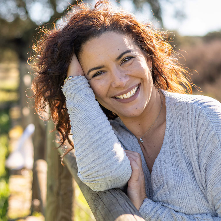 Smiling woman leaning on a wooden fence in a sunny outdoor setting.