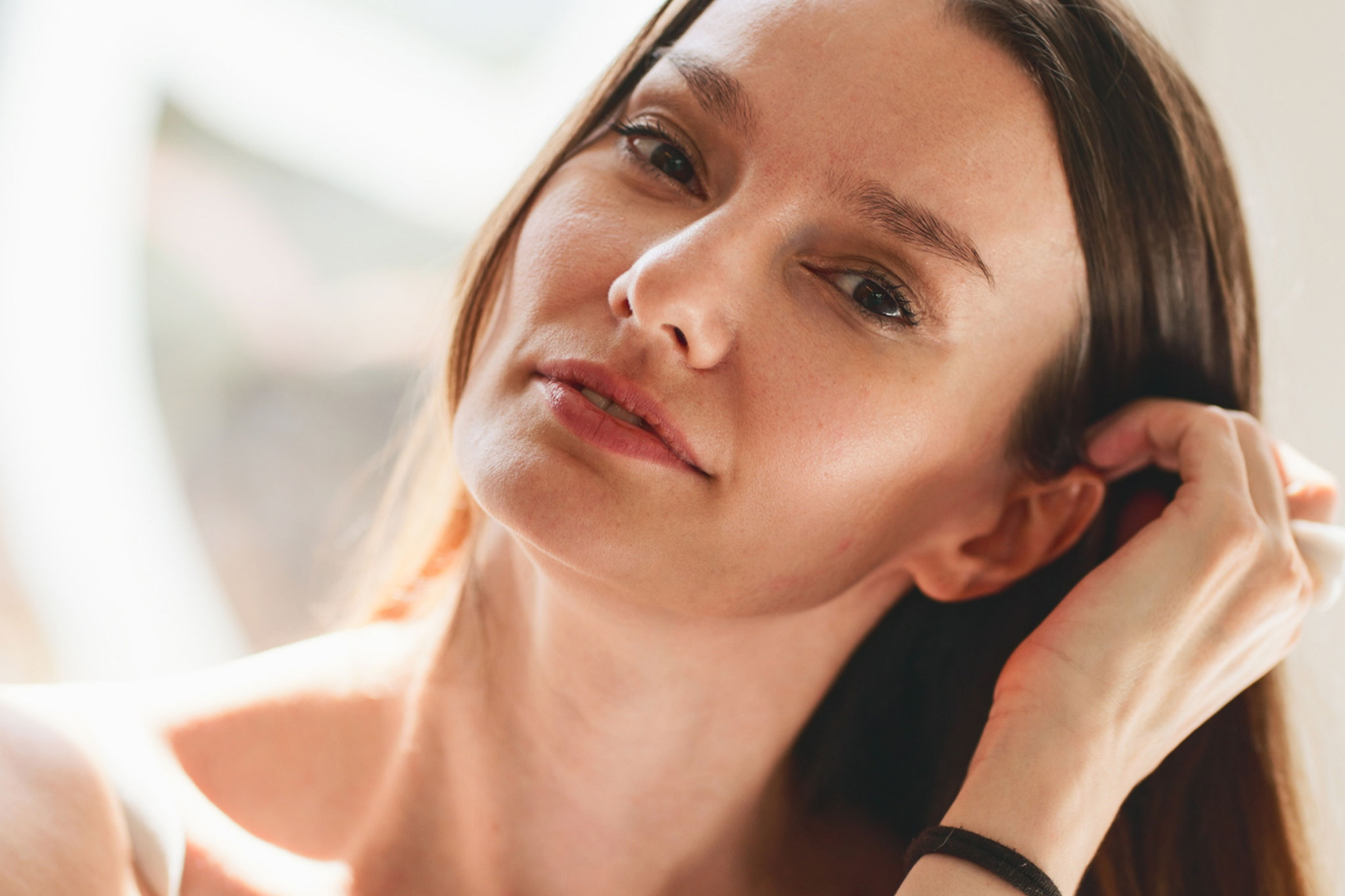A woman with long hair adjusts her hair, looking thoughtfully off to the side.