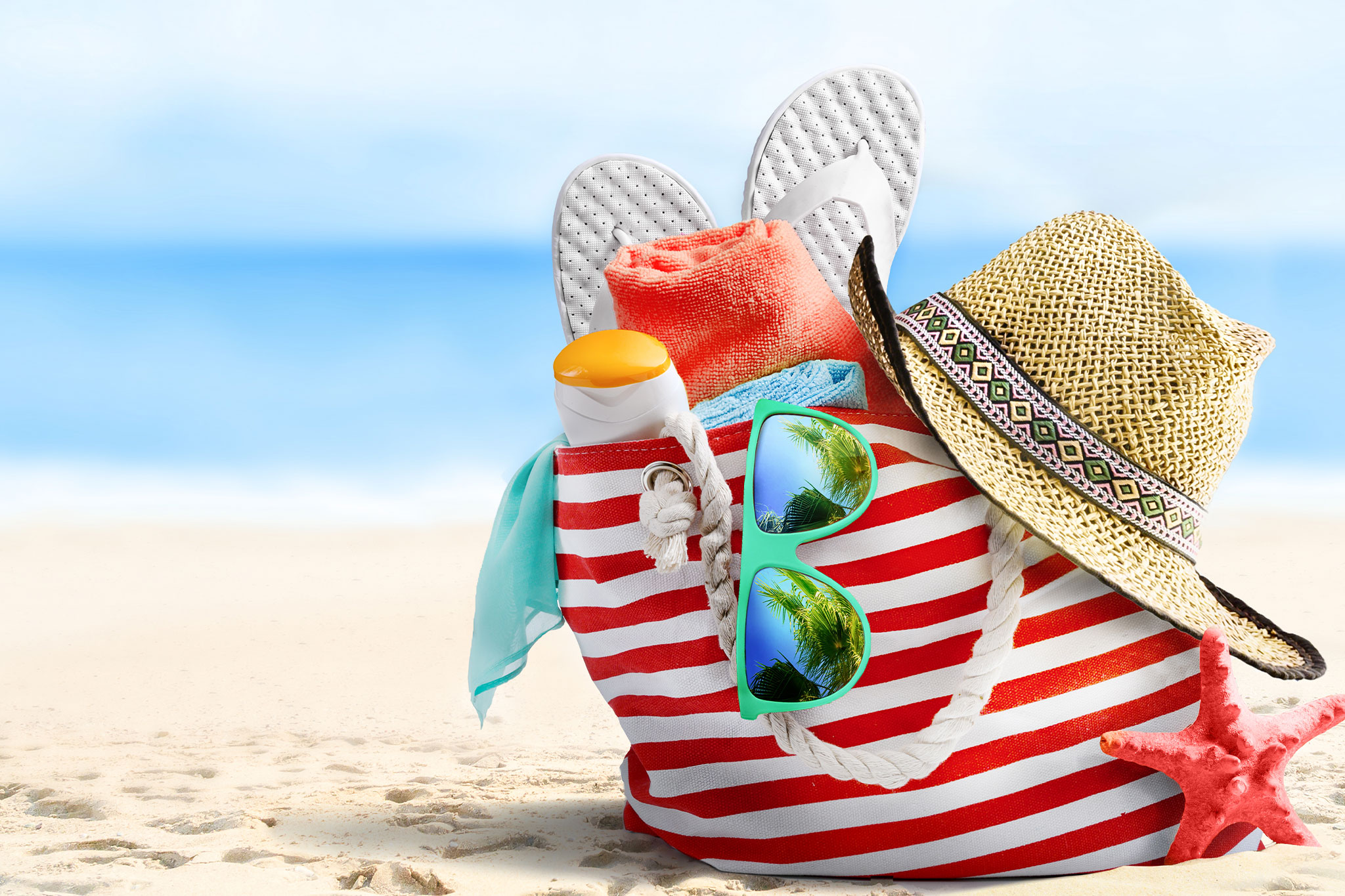Beach bag with sunglasses, straw hat, flip-flops, towel, and sunscreen on sandy beach background.
