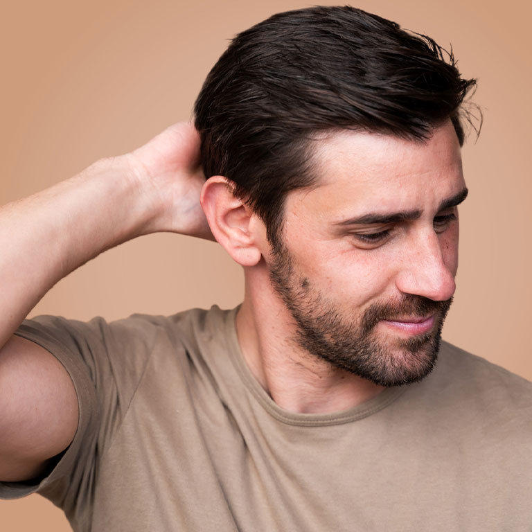 A person with short dark hair and a beard touches the back of their head, wearing a beige t-shirt.