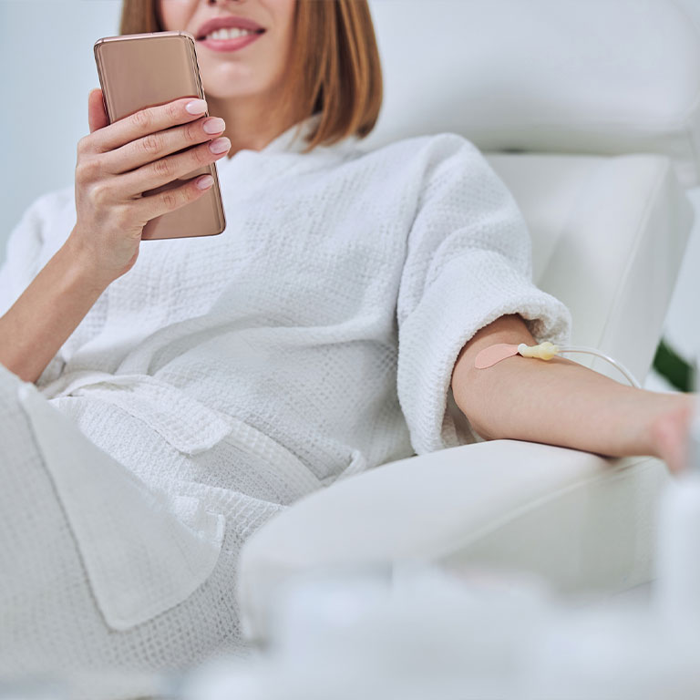 A woman in a white robe using a smartphone while receiving an IV treatment in a spa chair.