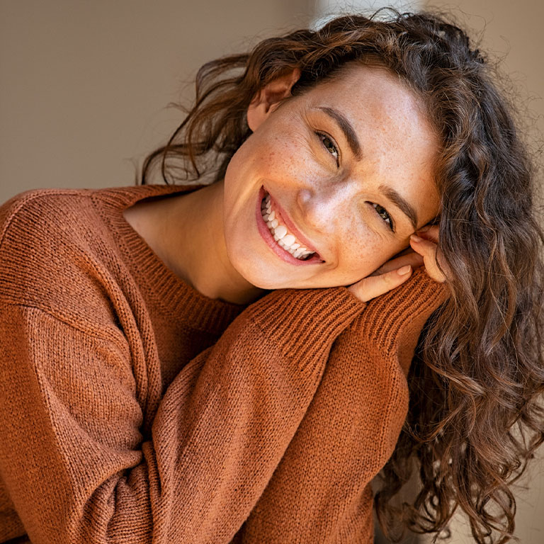 A woman with curly hair smiling while leaning on her hand, wearing a brown sweater.