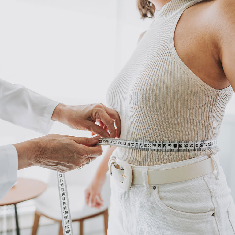 Person measuring waist with tape, wearing a sleeveless top, in a bright room.