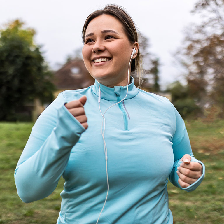 A woman jogging in a park, wearing a blue top and earbuds, smiling.