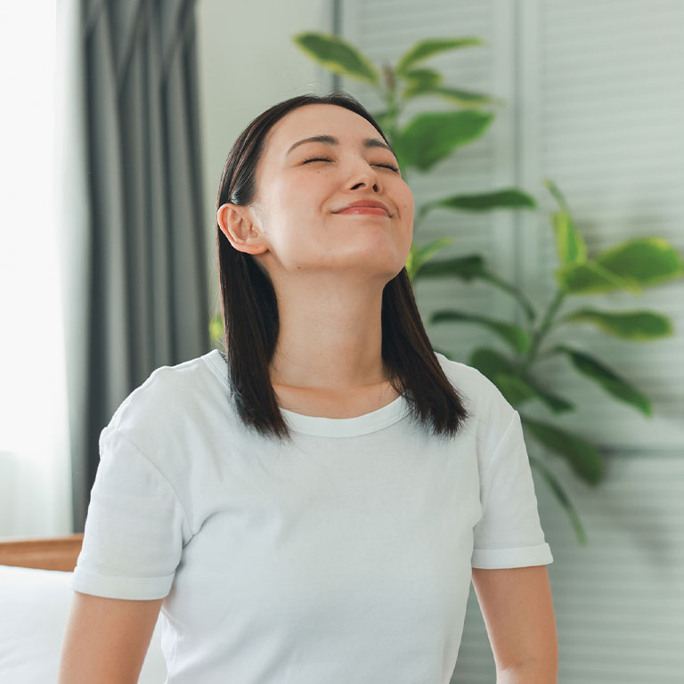 A woman in a white shirt relaxing with eyes closed near indoor plants.