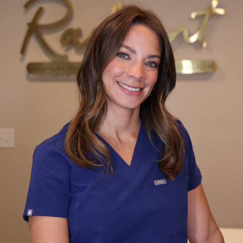 A woman in blue scrubs smiling in a medical office with blurred signage in the background.